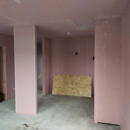 BWG- Big Work Group - Wnętrze w trakcie montażu płyt gipsowo-kartonowych na ścianach i suficie, widoczny surowy beton na podłodze, fragment okna PCV i płyta OSB oparta o ścianę.