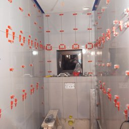 BWG- Big Work Group - Łazienka w trakcie układania szarych płytek, widoczne krzyżyki dystansowe i poziomica, małe okno w głębi.