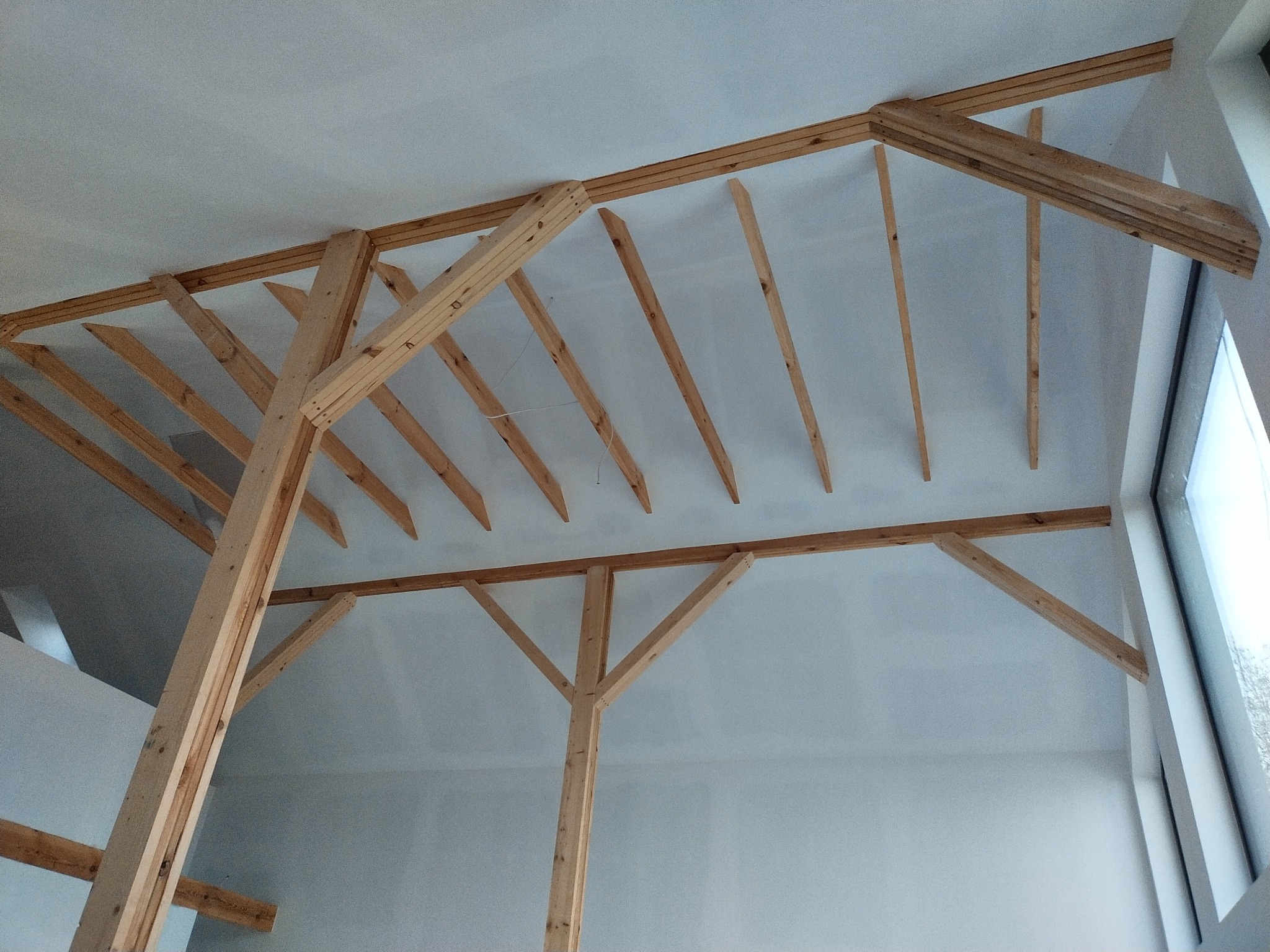 Drewniana konstrukcja szkieletowa dachu w surowym stanie, widoczna wewnątrz jasnego pomieszczenia z oknem.