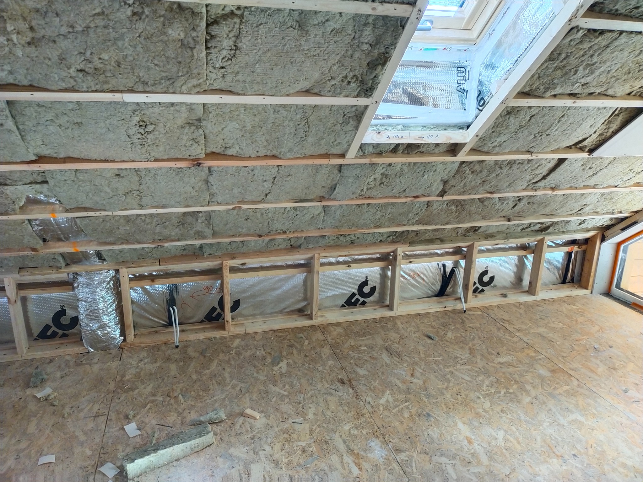 Poddasze w trakcie ocieplania wełną mineralną, widoczne drewniane legary, folia aluminiowa oraz fragment okna dachowego.