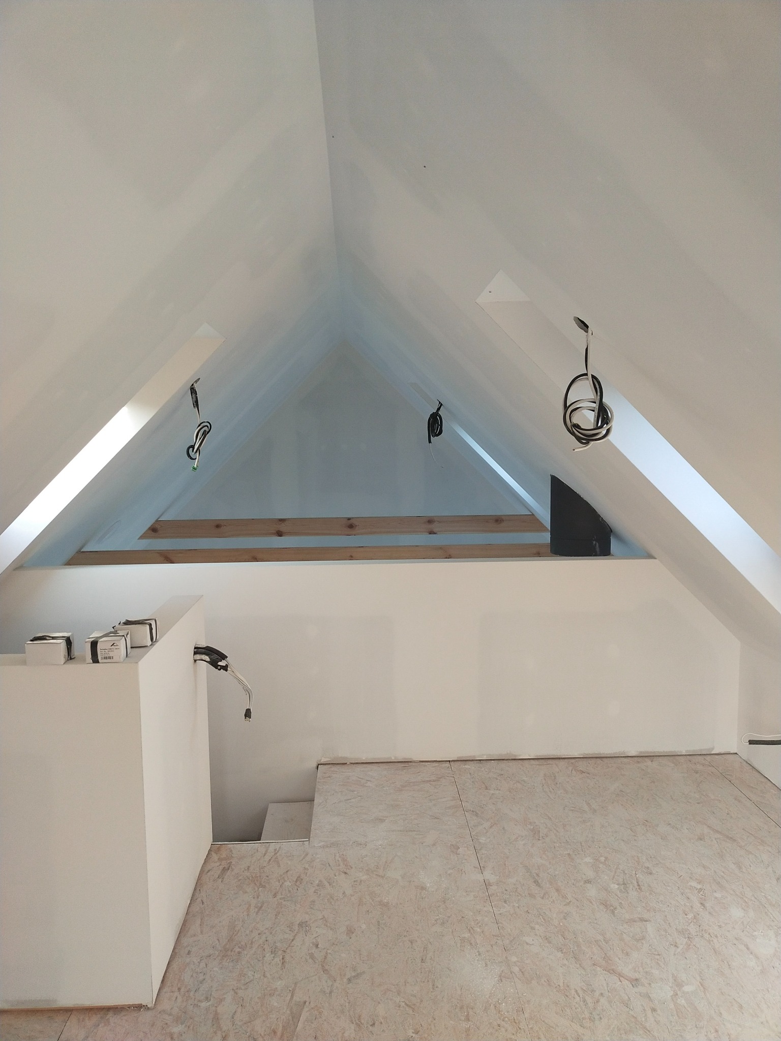 Poddasze w trakcie adaptacji: białe ściany szczytowe, drewniana konstrukcja więźby dachowej, surowa podłoga z płyt OSB, zwisające przewody elektryczne i kartony z osprzętem.