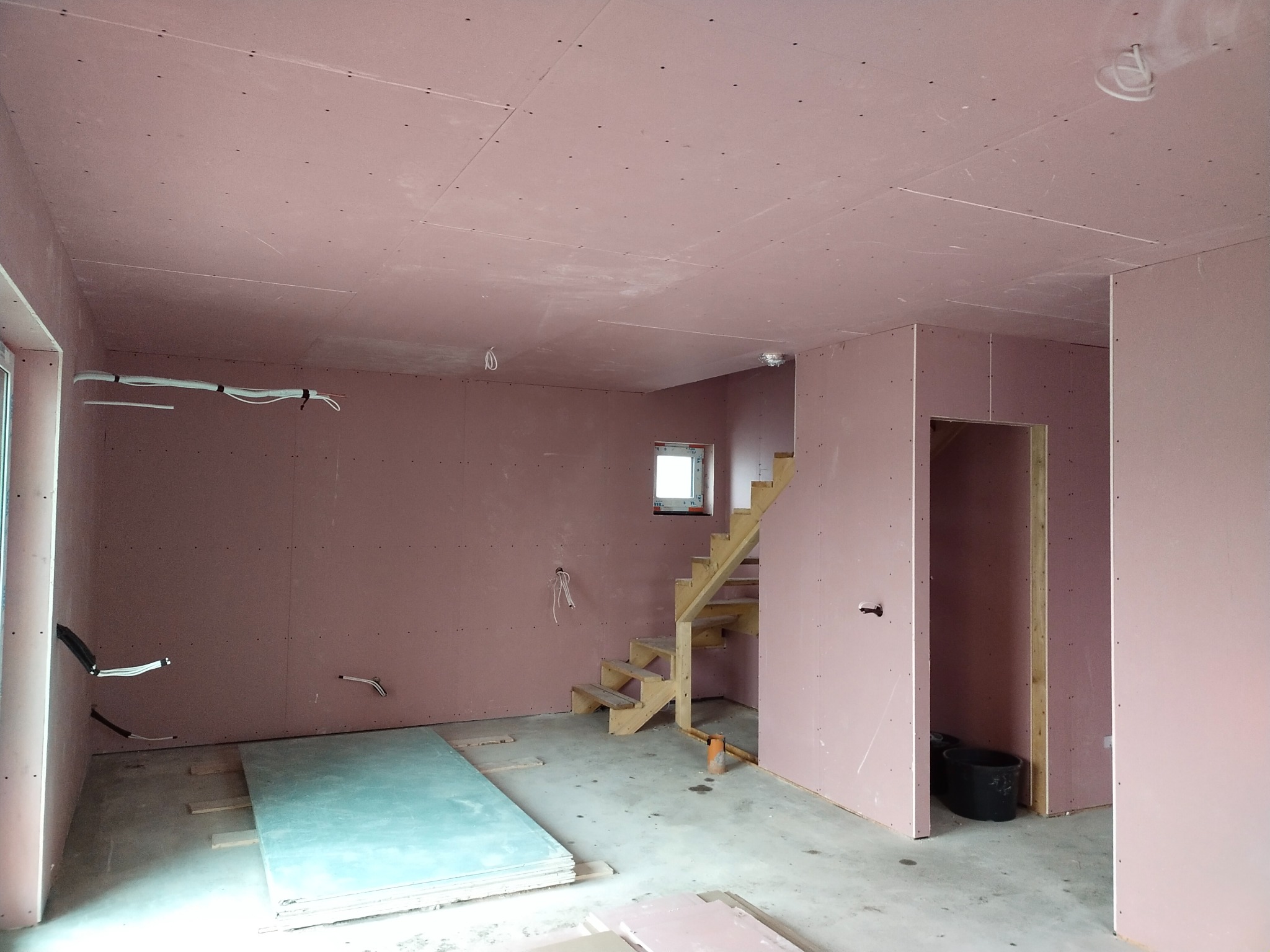 Surowe wnętrze z płyt gipsowo-kartonowych w odcieniach różu, widoczne drewniane schody, okno, instalacje elektryczne i stos płyt na podłodze.