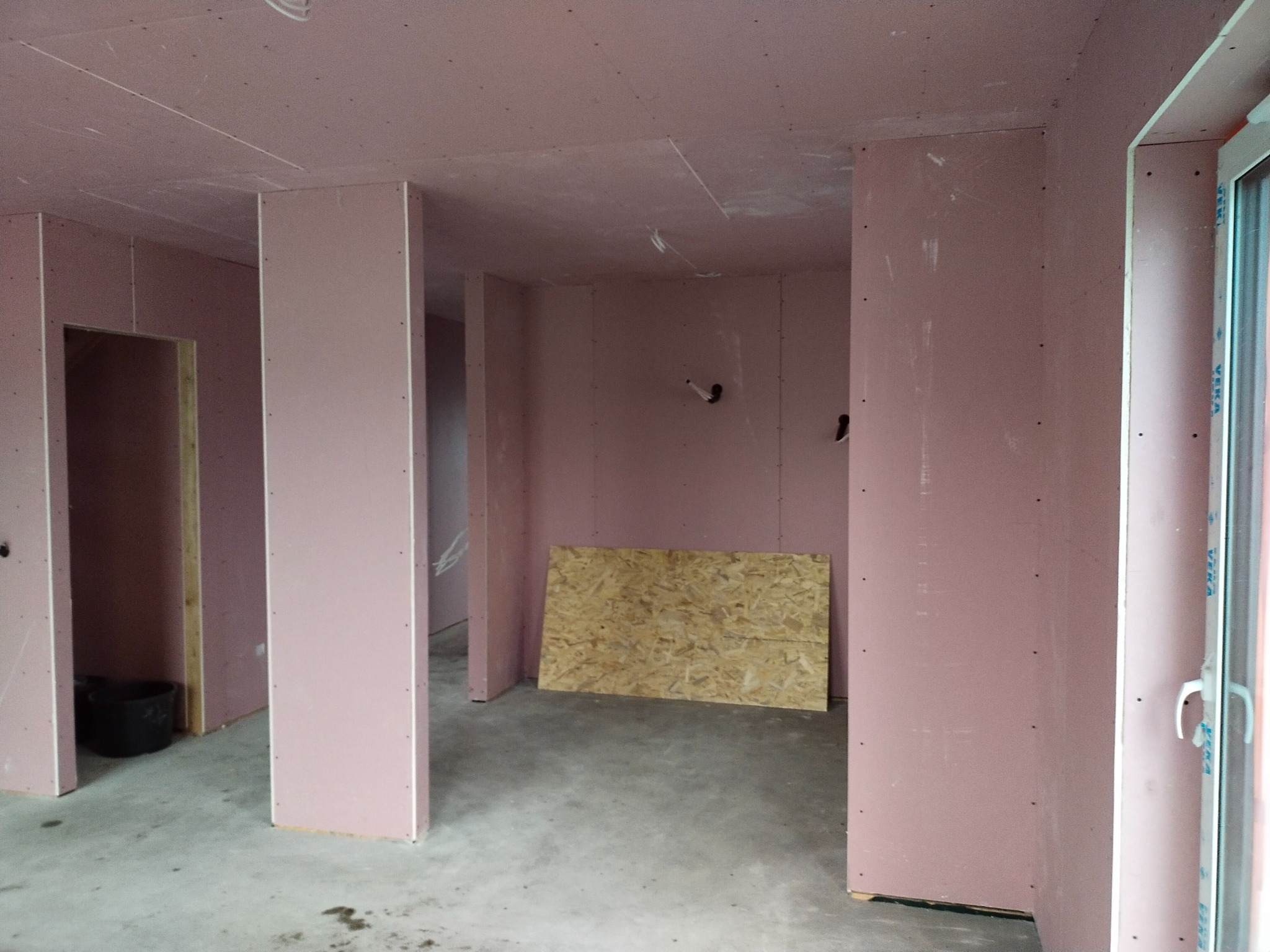 Wnętrze w trakcie montażu płyt gipsowo-kartonowych na ścianach i suficie, widoczny surowy beton na podłodze, fragment okna PCV i płyta OSB oparta o ścianę.