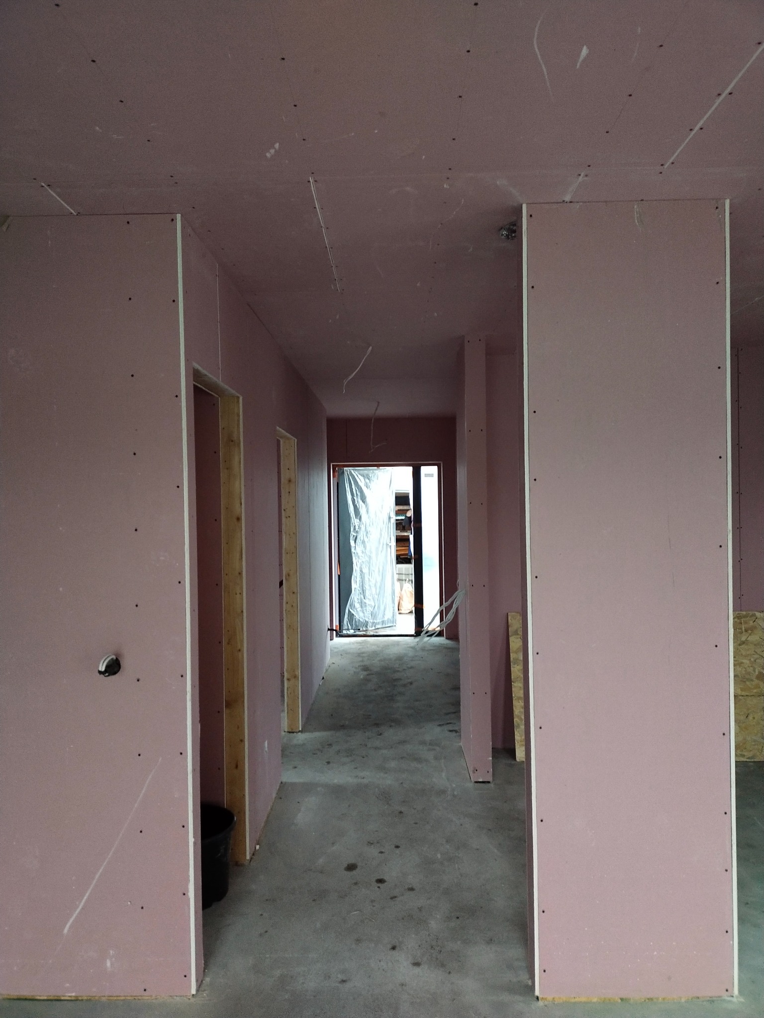 Korytarz w trakcie montażu płyt gipsowo-kartonowych o różowym zabarwieniu, widoczne drewniane ościeżnice drzwiowe i surowy beton na podłodze.