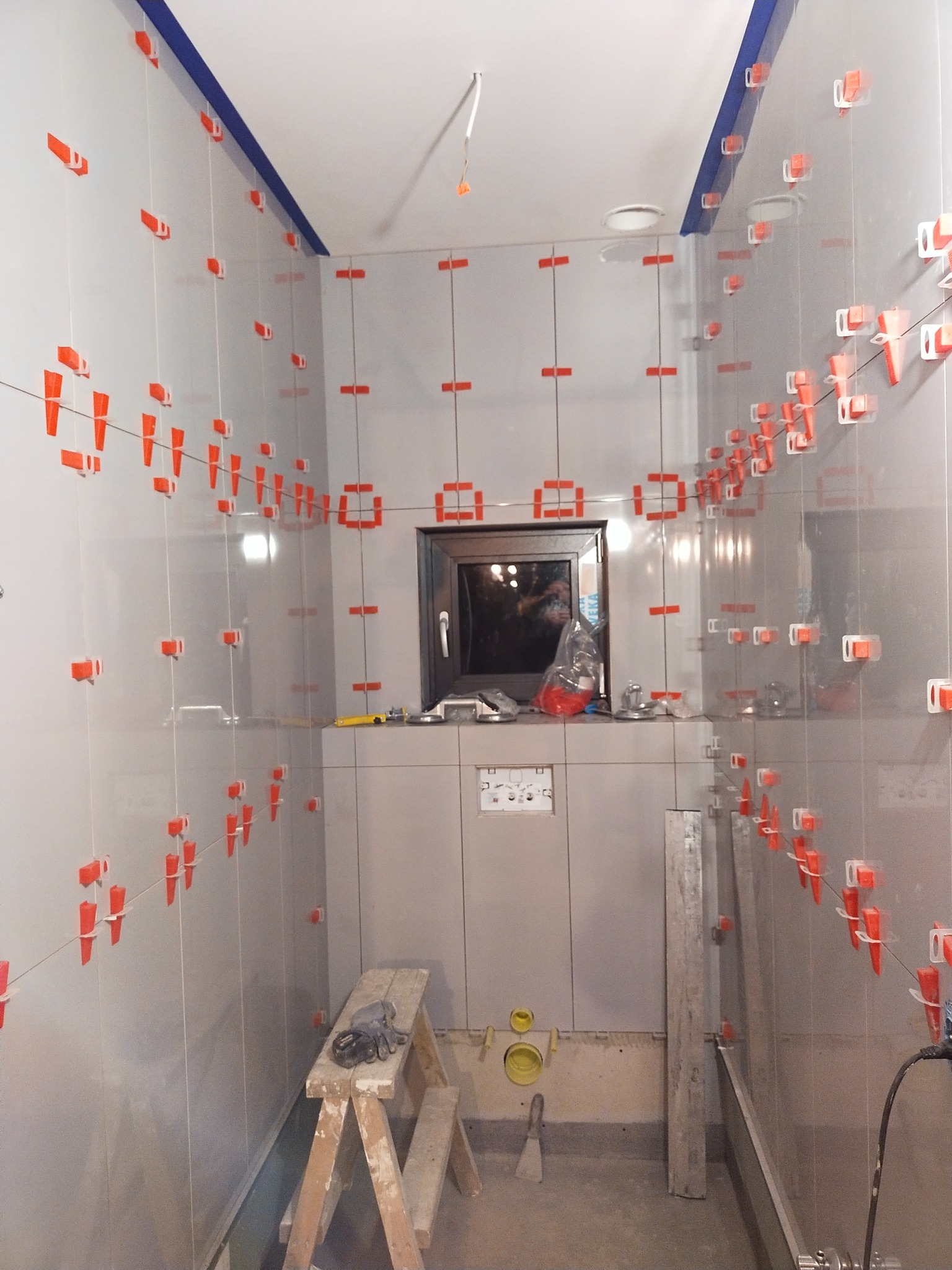 Łazienka w trakcie układania szarych płytek, widoczne krzyżyki dystansowe i poziomica, małe okno w głębi.