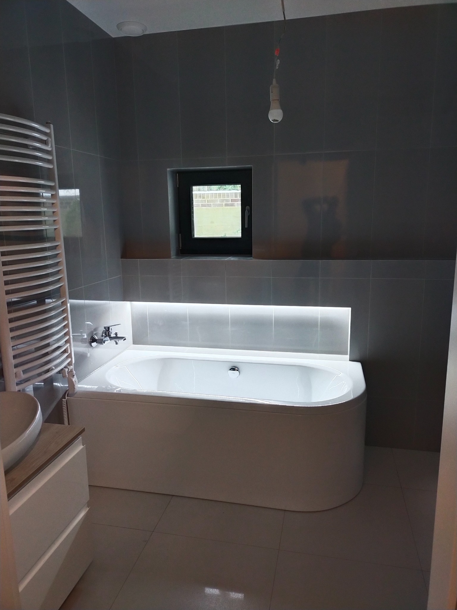 Nowoczesna łazienka z szarymi płytkami, białą wanną podświetloną ledami, grzejnikiem drabinkowym i wiszącą żarówką na kablu.