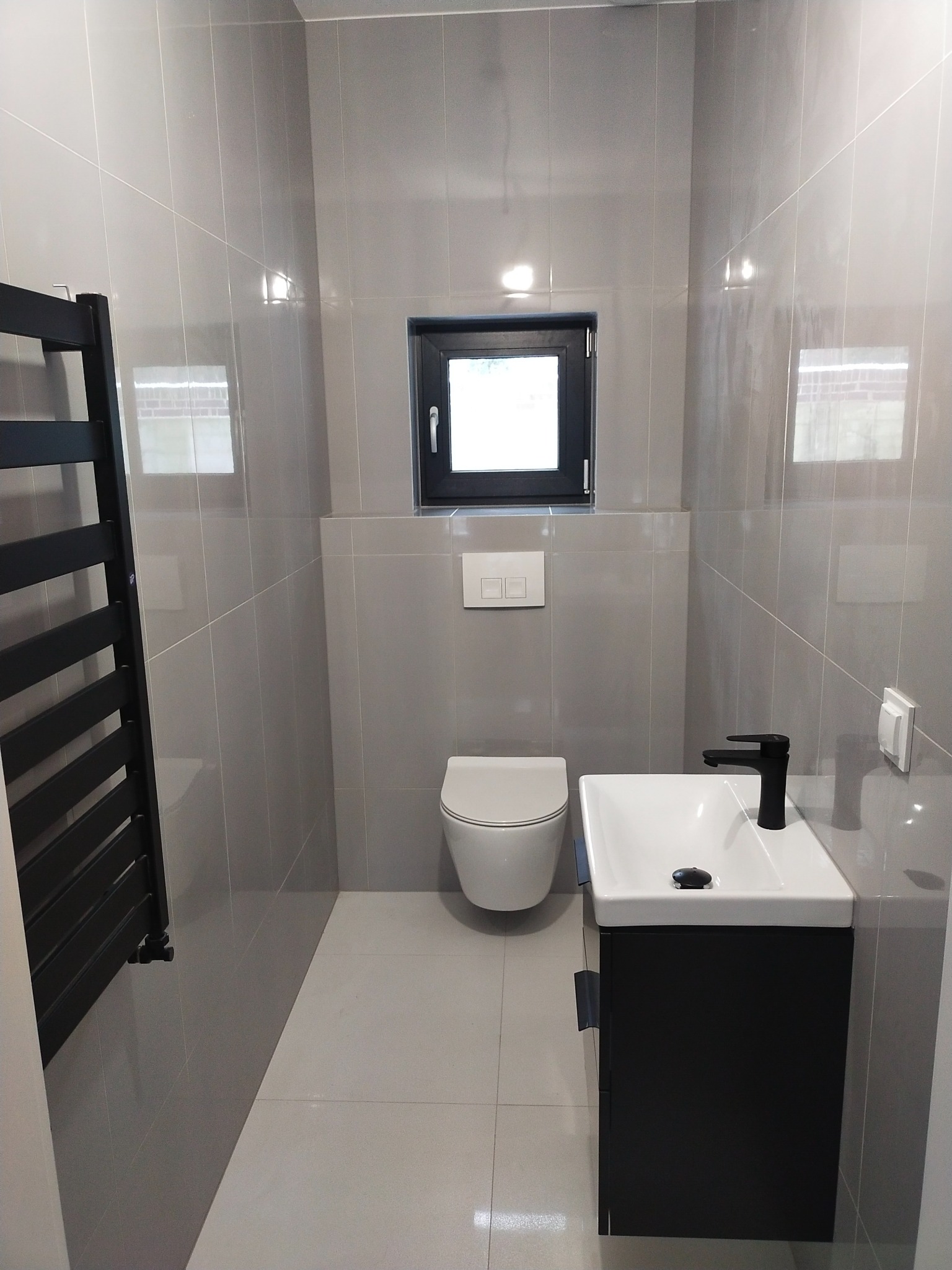 Wykończona łazienka w odcieniach szarości z czarnymi akcentami: grzejnik drabinkowy, bateria umywalkowa i rama okienna. Umywalka nablatowa z szafką, podwieszana toaleta. Widoczne płytki ceramiczne...