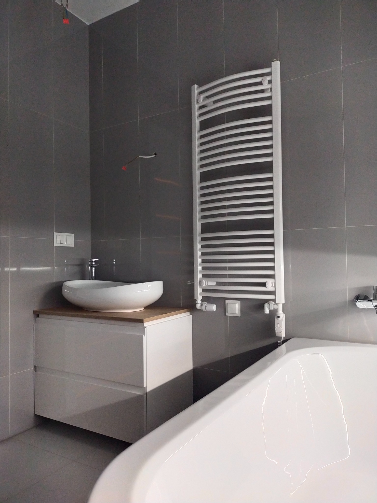 Nowoczesna łazienka w Piasecznie z szarymi płytkami, białą umywalką nablatową na szafce z drewnianym blatem, białym grzejnikiem drabinkowym i fragmentem wanny.