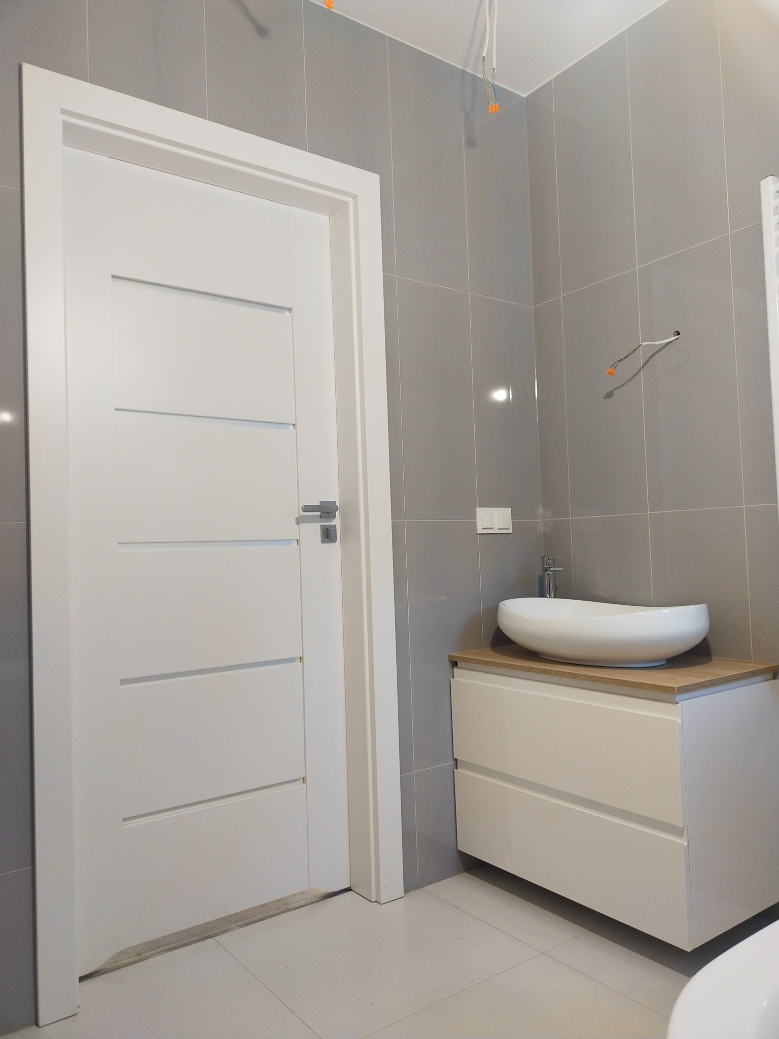 Nowoczesna łazienka z szarymi płytkami, białą szafką pod umywalkę z blatem imitującym drewno i białą umywalką nablatową o owalnym kształcie, widoczna biała, minimalistyczna ościeżnica drzwiowa.