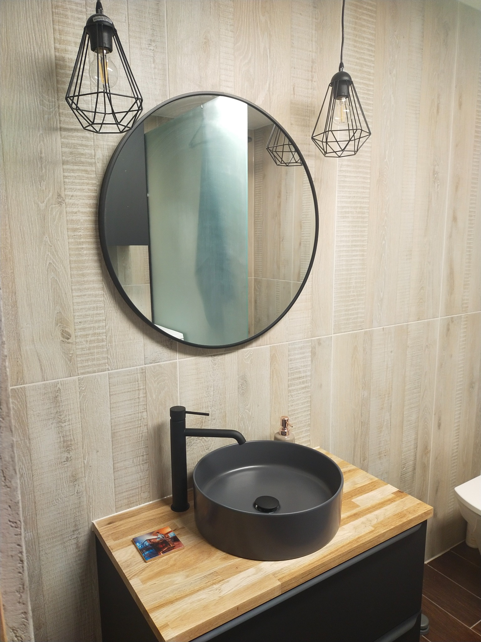 Nowoczesna łazienka z okrągłym lustrem w czarnej ramie, umywalką w kolorze grafitowym na drewnianym blacie i lampami wiszącymi w geometrycznych kloszach z czarnego drutu, wykończona płytkami...