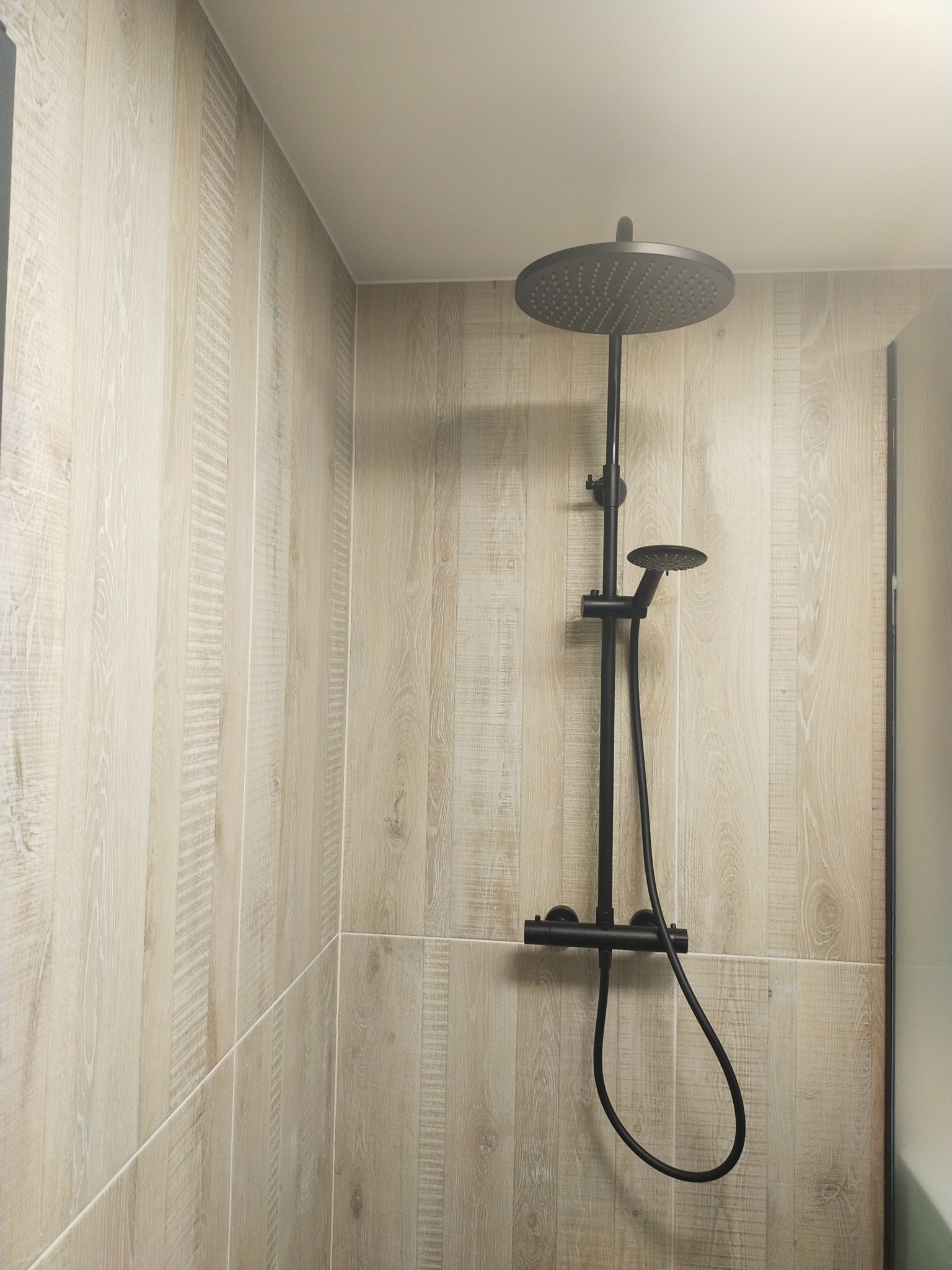 Nowoczesny, czarny zestaw prysznicowy z deszczownicą, zamontowany na ścianie wyłożonej płytkami imitującymi jasne drewno w kabinie prysznicowej.