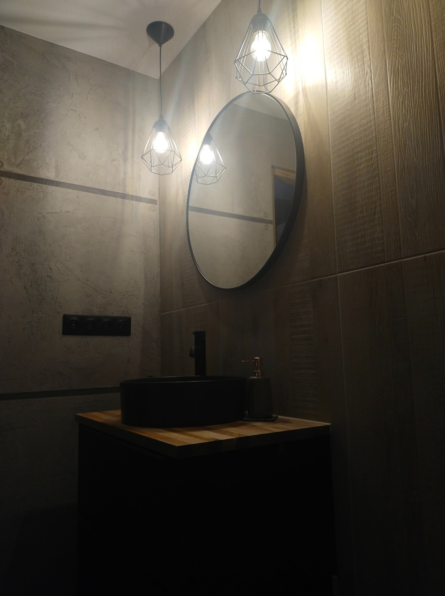 Nowoczesna łazienka z okrągłym lustrem w czarnej ramie, czarną umywalką na drewnianym blacie i designerskimi lampami wiszącymi w metalowych klatkach, podkreślona płytkami imitującymi drewno...