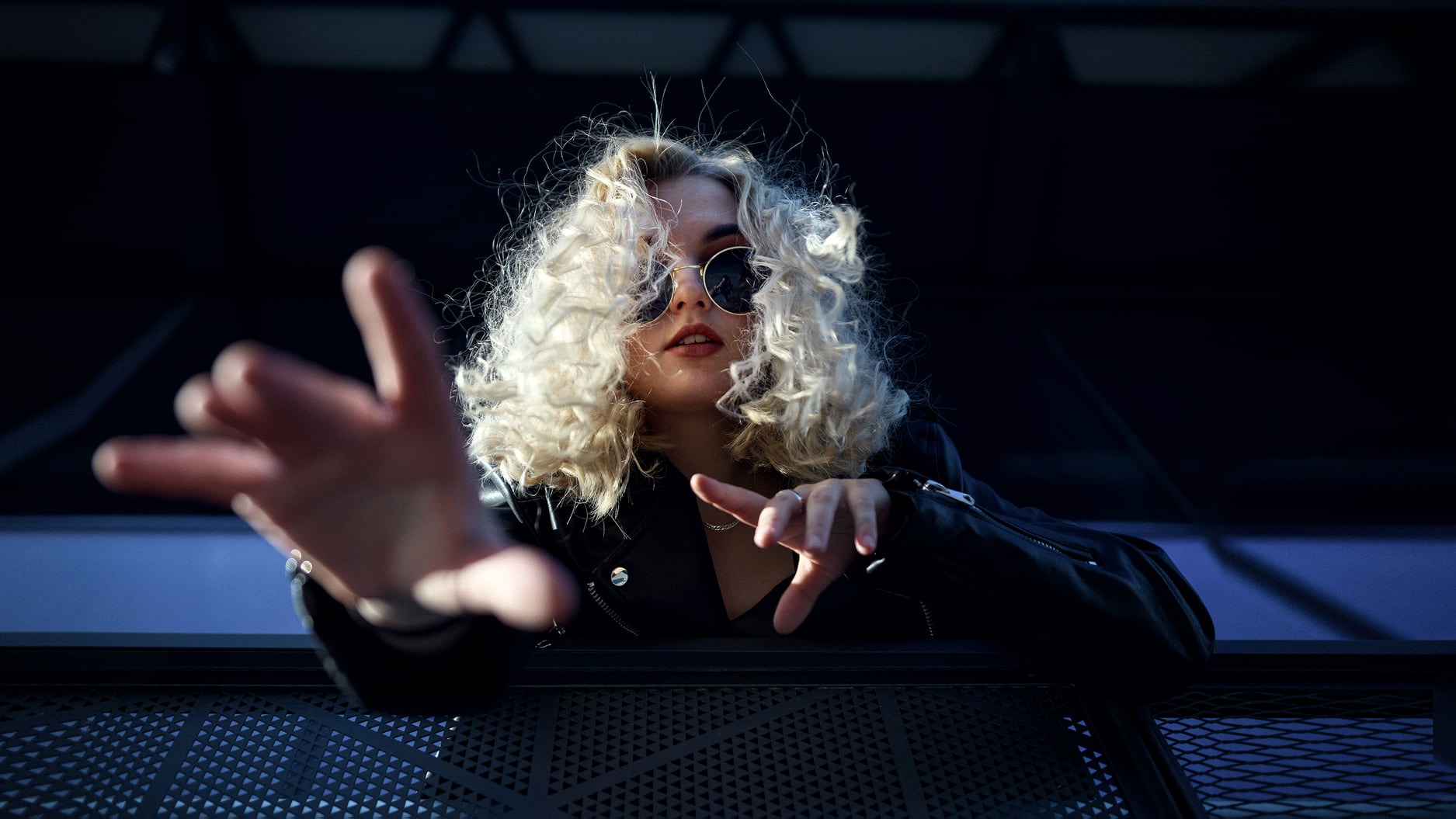 Portret kobiety z blond kręconymi włosami i okularami przeciwsłonecznymi, w czarnej skórzanej kurtce, wyciągającej dłoń w stronę obiektywu, na tle ciemnego, geometrycznego tła.