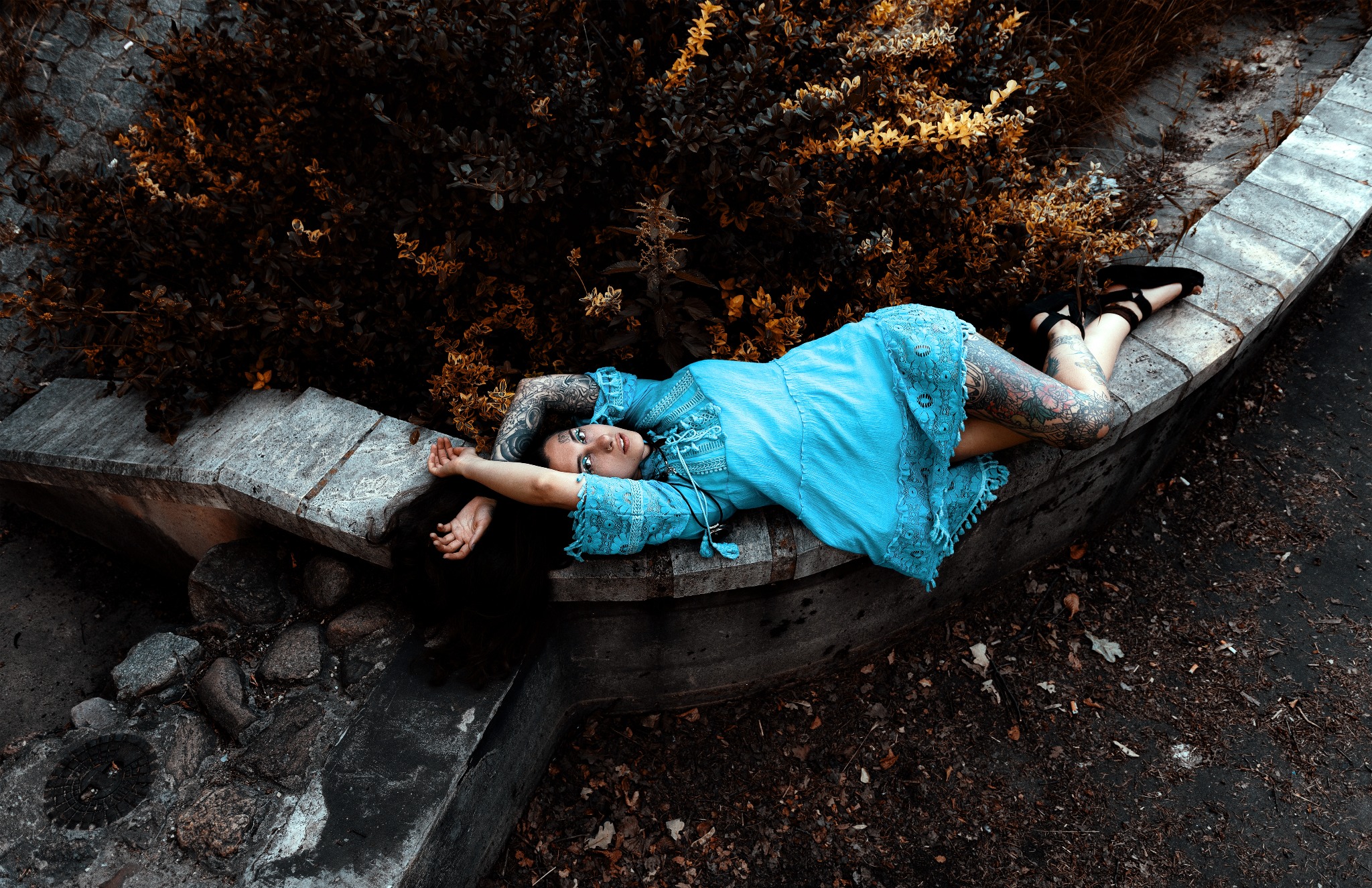 Kobieta z tatuażami w błękitnej sukience leży na betonowym murku, otoczona roślinnością o jesiennych barwach.