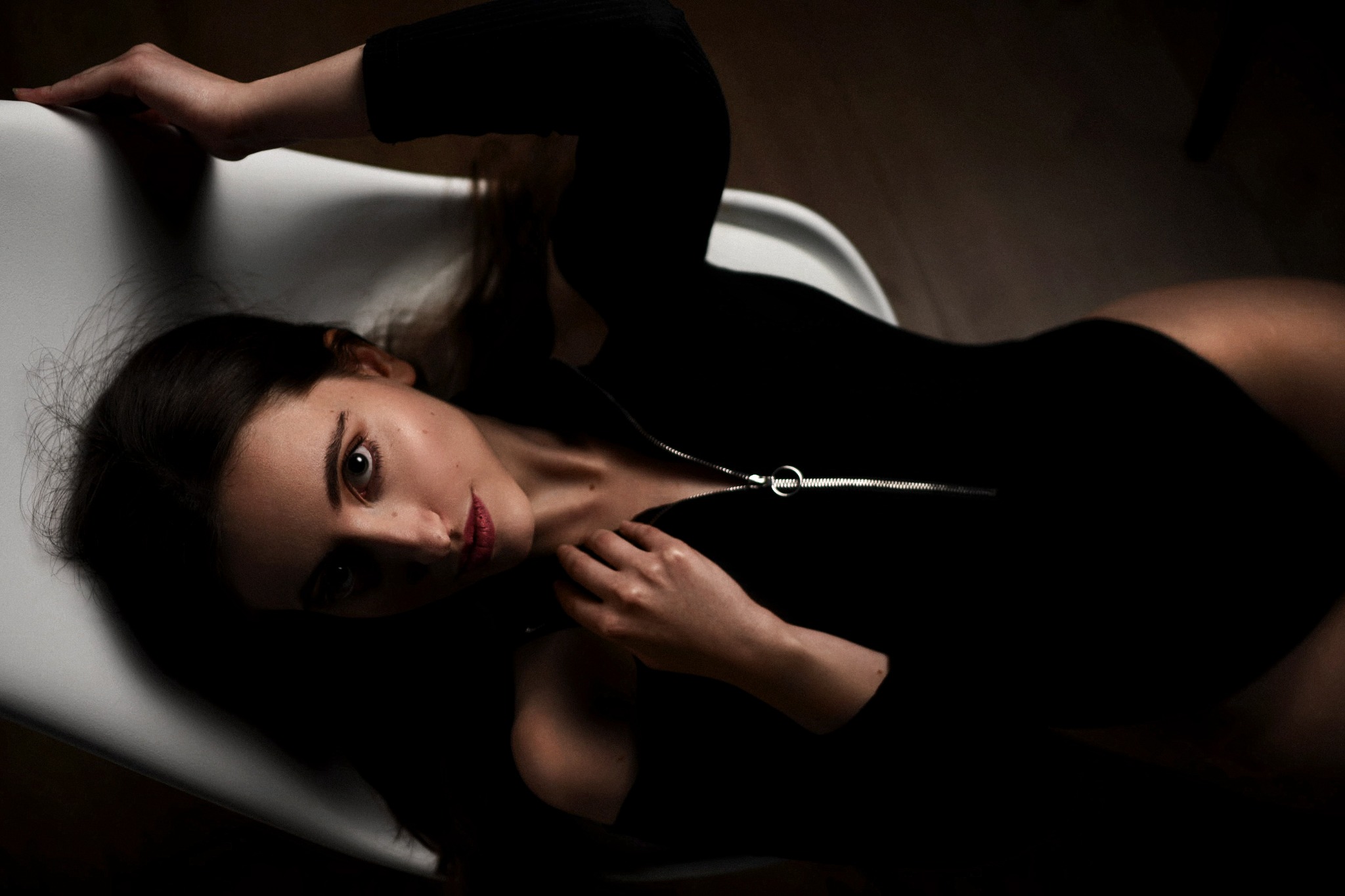 Portret kobiety w czarnym body z suwakiem, leżącej na białym krześle, z głową skierowaną w stronę obiektywu, w ciemnym otoczeniu.