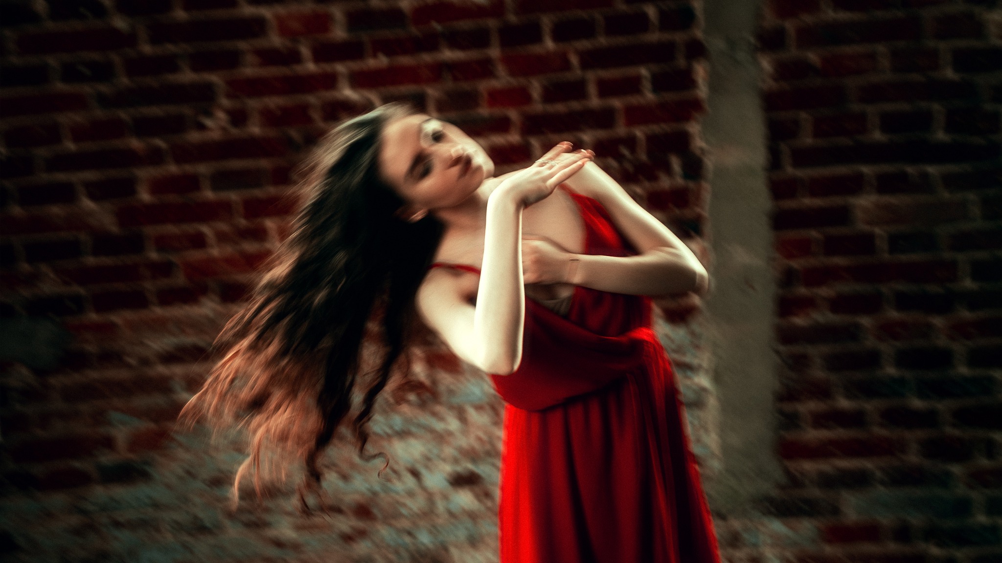 Kobieta w czerwonej sukience, z rozwianymi włosami, pozująca na tle ceglanej ściany w dynamicznej pozie.
