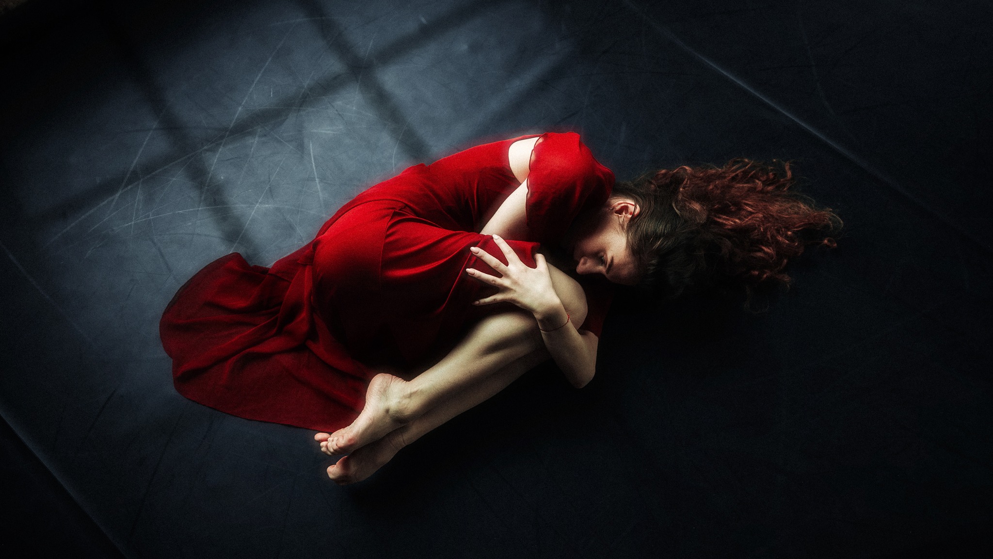 Kobieta skulona na ciemnym tle w czerwonej, zwiewnej sukni, z rozpuszczonymi, rudymi włosami i delikatnym oświetleniem padającym z góry.