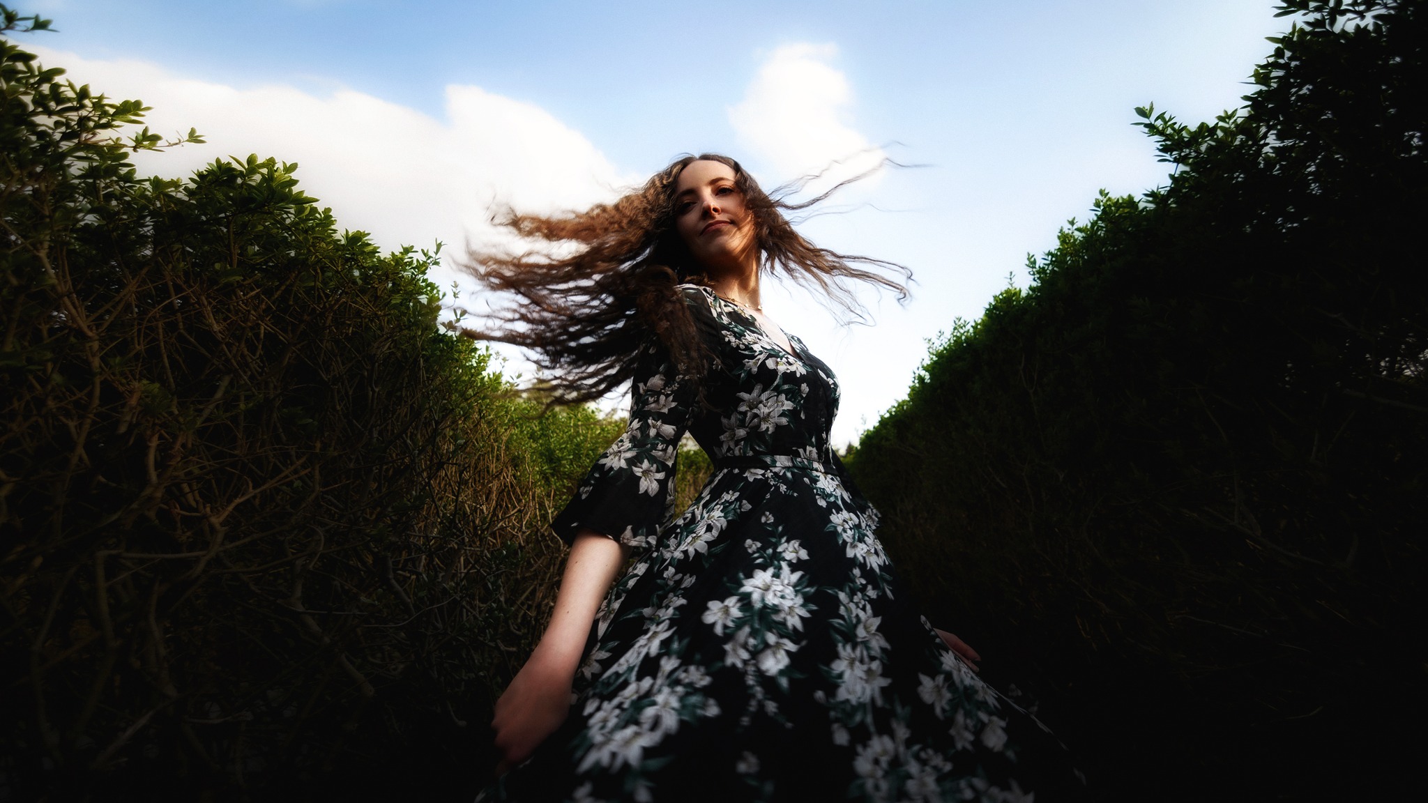 Kobieta w kwiecistej sukience pozuje w labiryncie z żywopłotu, włosy rozwiane na wietrze, ujęcie z dołu.