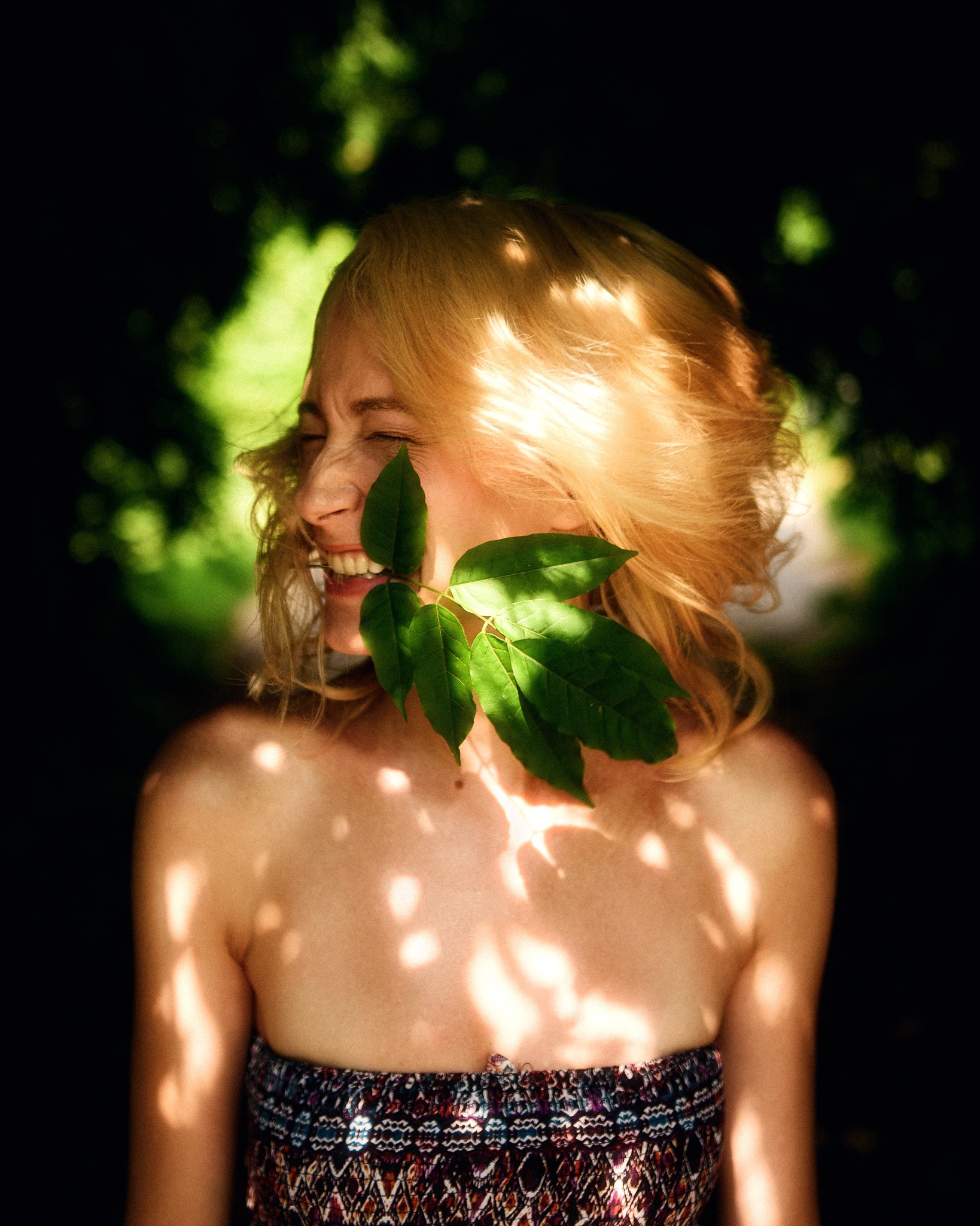Uśmiechnięta blondynka z liściem w ustach, oświetlona plamami światła słonecznego w leśnym otoczeniu.