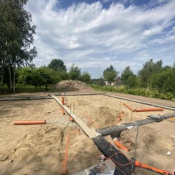 Przedsiębiorstwo Handlowo Usługowe DOLA DANIEL DOBROWOLSKI - Fundamenty domu w trakcie budowy, widoczne rury kanalizacyjne, poziomica i narzędzia na tle zieleni i błękitnego nieba z chmurami.