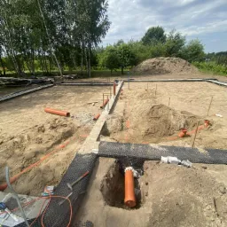 Fundament w trakcie budowy z widocznymi rurami kanalizacyjnymi, poziomica i narzędzia. W tle drzewa i usypana ziemia.