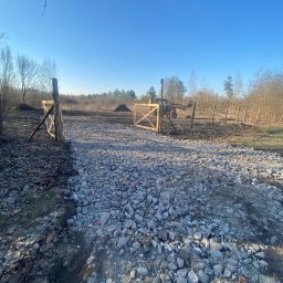 Przedsiębiorstwo Handlowo Usługowe DOLA DANIEL DOBROWOLSKI - Ułożona droga z kamieni prowadząca przez drewnianą bramę na tle pola i lasu w słoneczny dzień. Widoczna maszyna budowlana w tle.