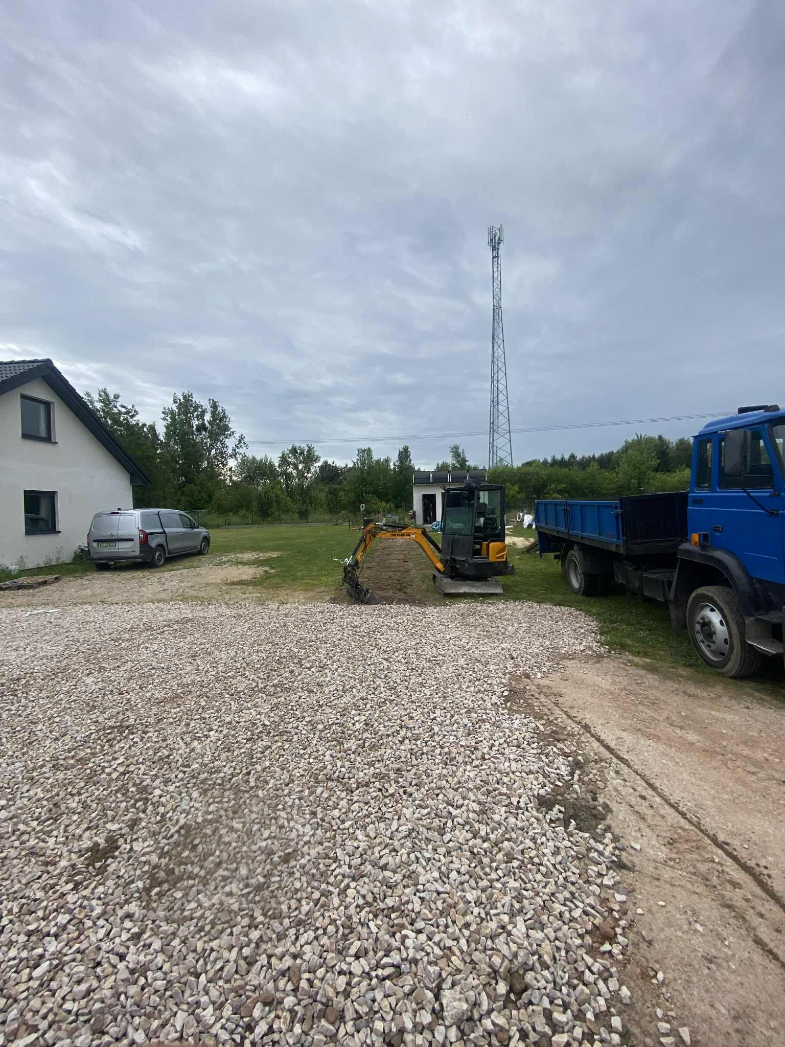 Mini koparka Hyundai na żwirowym podłożu, obok niebieska ciężarówka. W tle dom, trawnik i wieża telekomunikacyjna. Prace ziemne w toku.