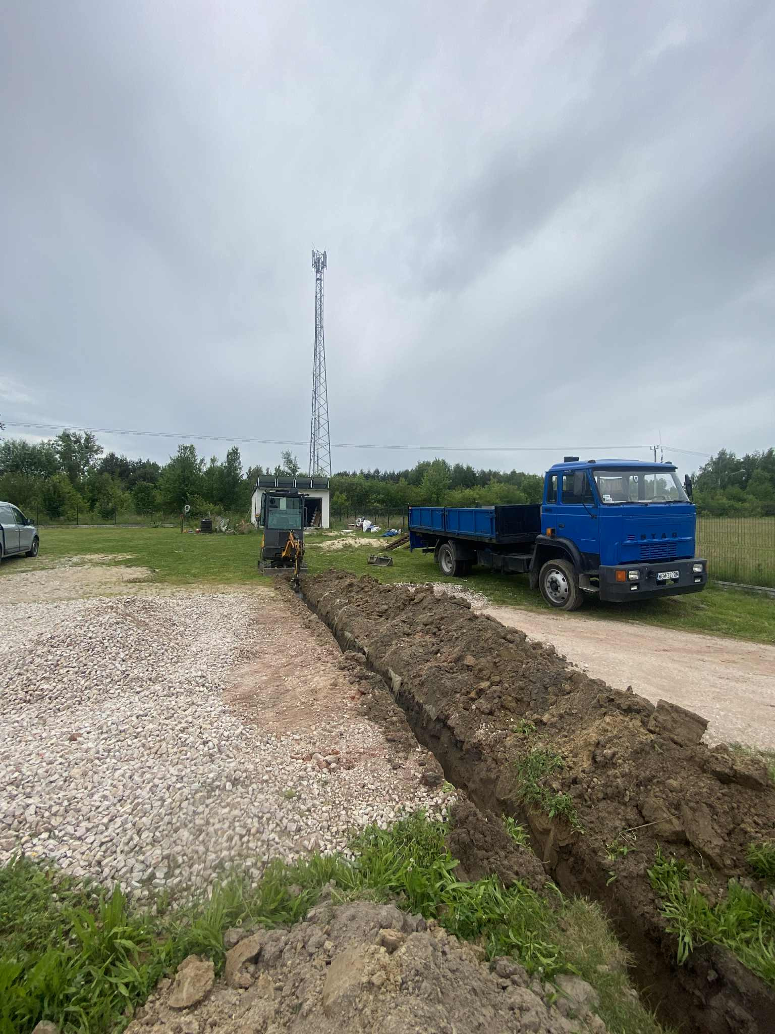 Wykopany rów na działce z gruzem, mała koparka obok, w tle niebieska ciężarówka i wieża telekomunikacyjna pod pochmurnym niebem.