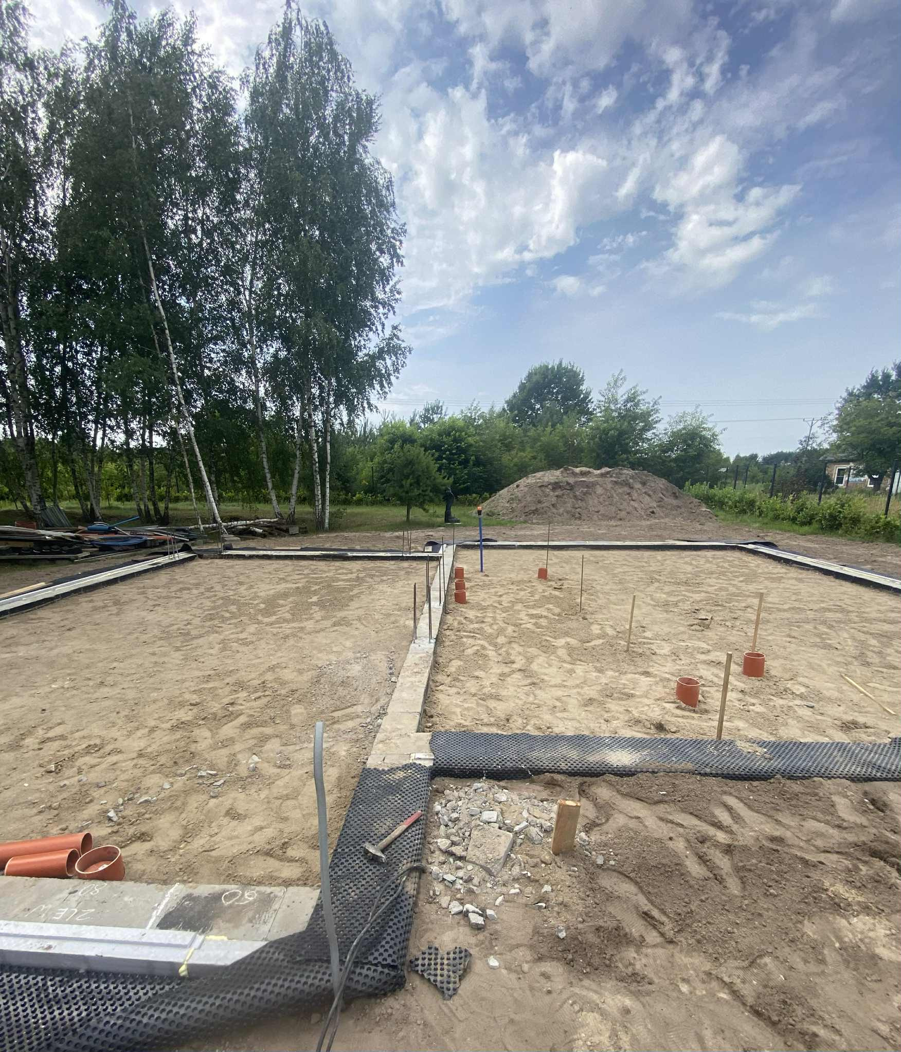 Fundamenty domu w trakcie budowy, widoczne szalunki, izolacja, rury kanalizacyjne i teren przygotowany pod wylewkę. W tle drzewa i niebo.