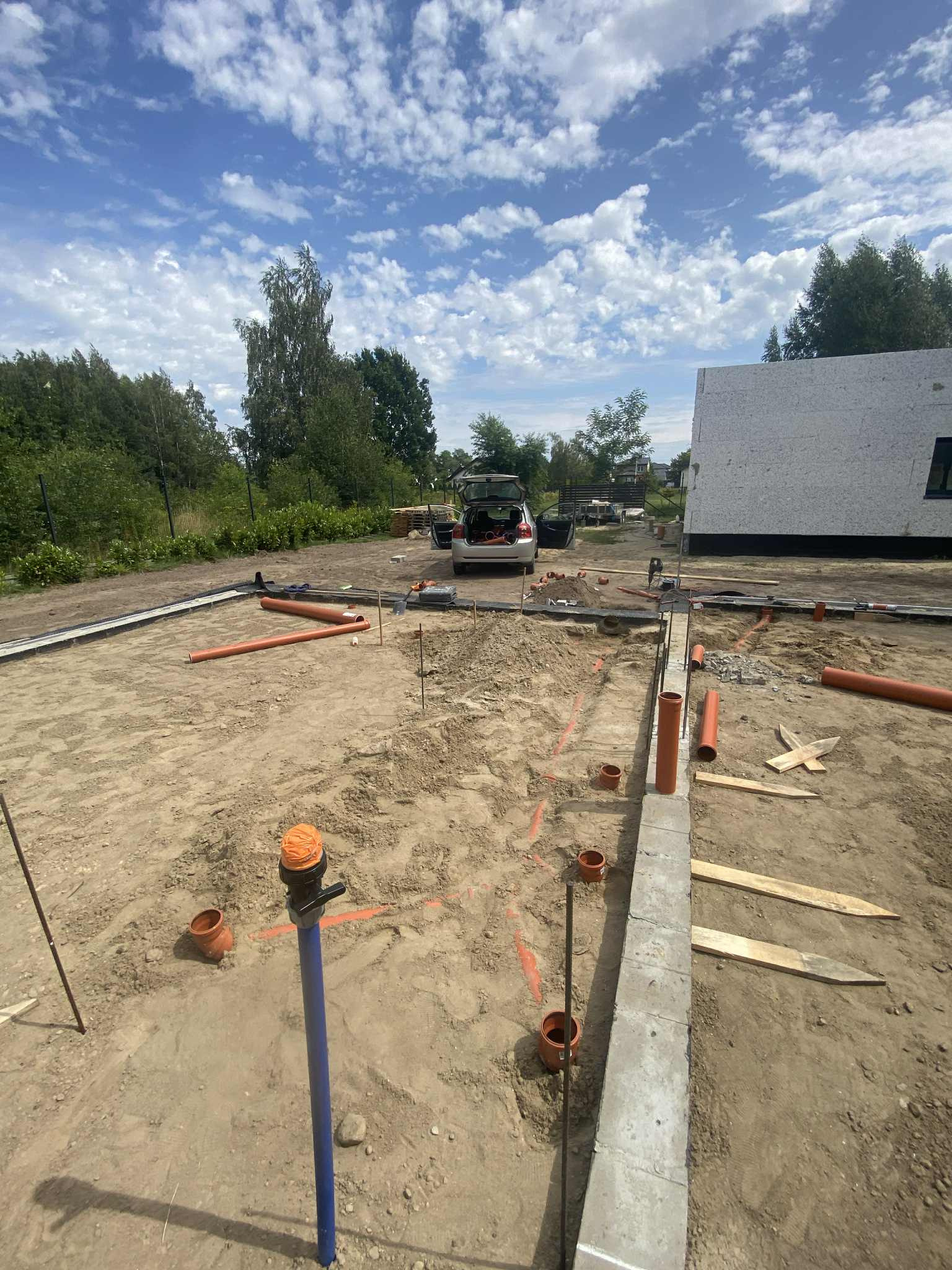 Instalacja rur kanalizacyjnych na placu budowy, widoczne rury pomarańczowe i niebieskie, częściowo ułożone w wykopie, w tle budynek w stanie surowym otwartym.