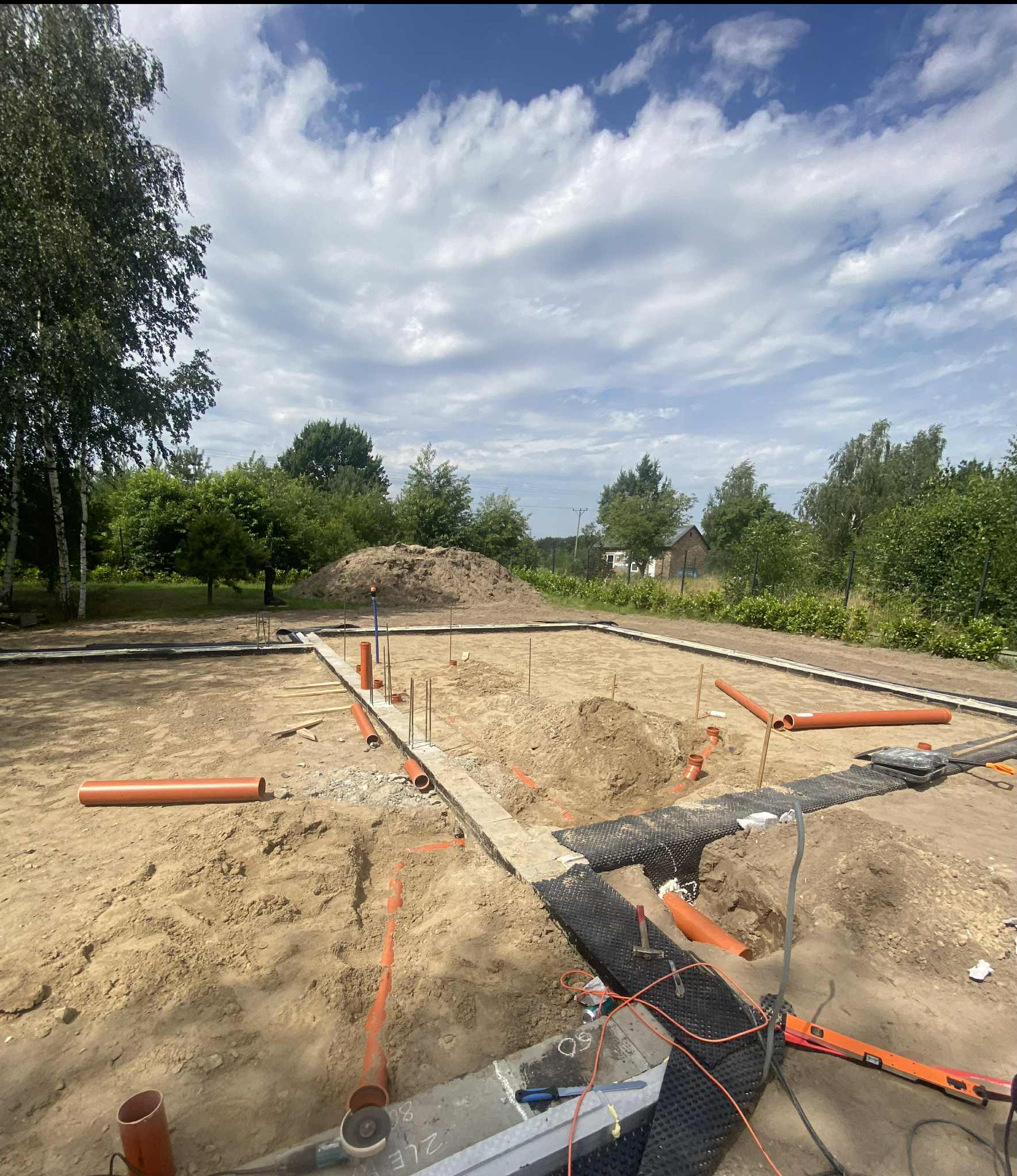 Fundamenty domu w trakcie budowy, widoczne rury kanalizacyjne, poziomica i narzędzia na tle zieleni i błękitnego nieba z chmurami.