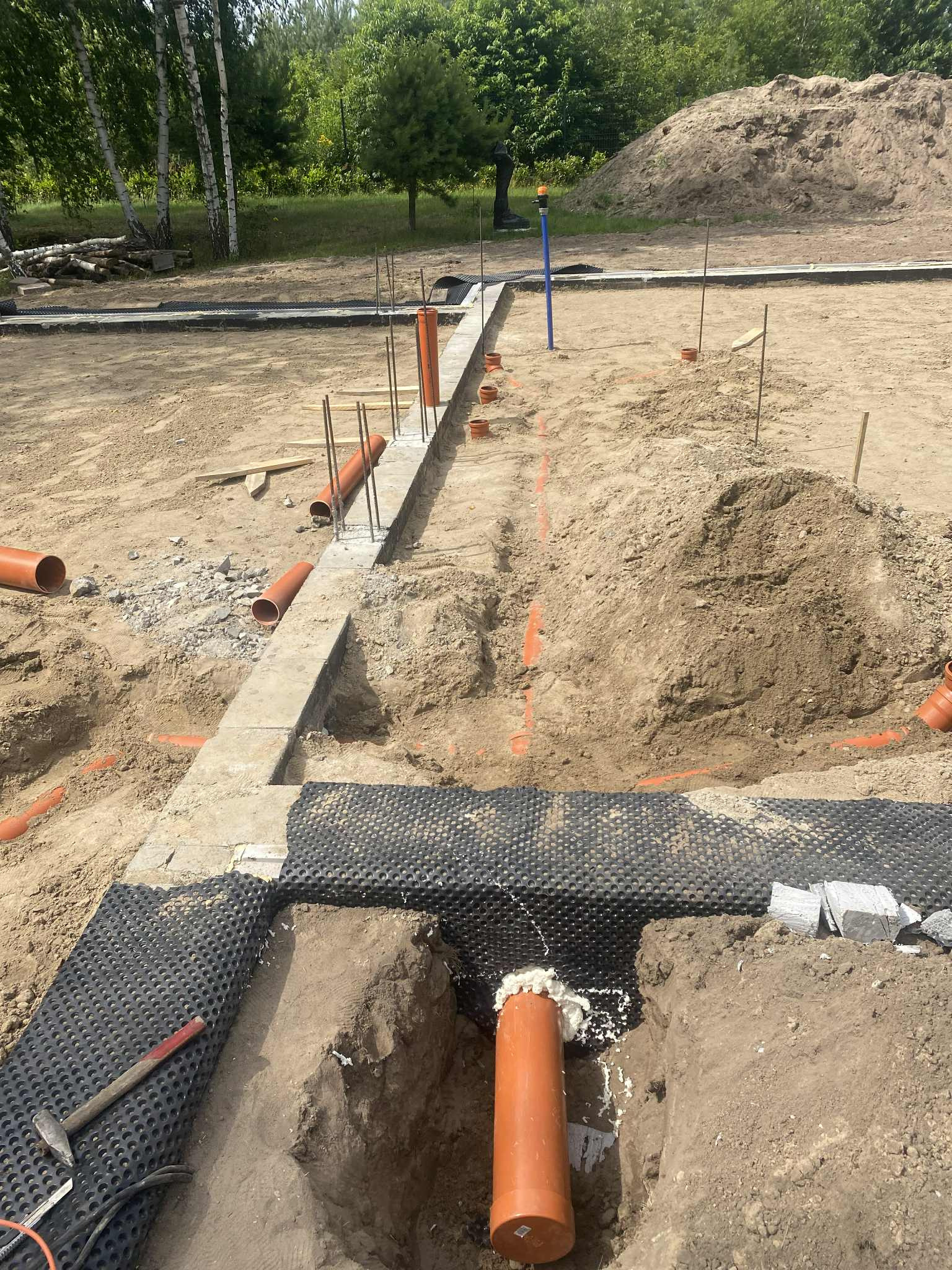 Fundamenty domu w trakcie budowy: widoczne rury kanalizacyjne, zbrojenie i czarna folia izolacyjna. Wykopane rowy w ziemi, przygotowane do dalszych prac.