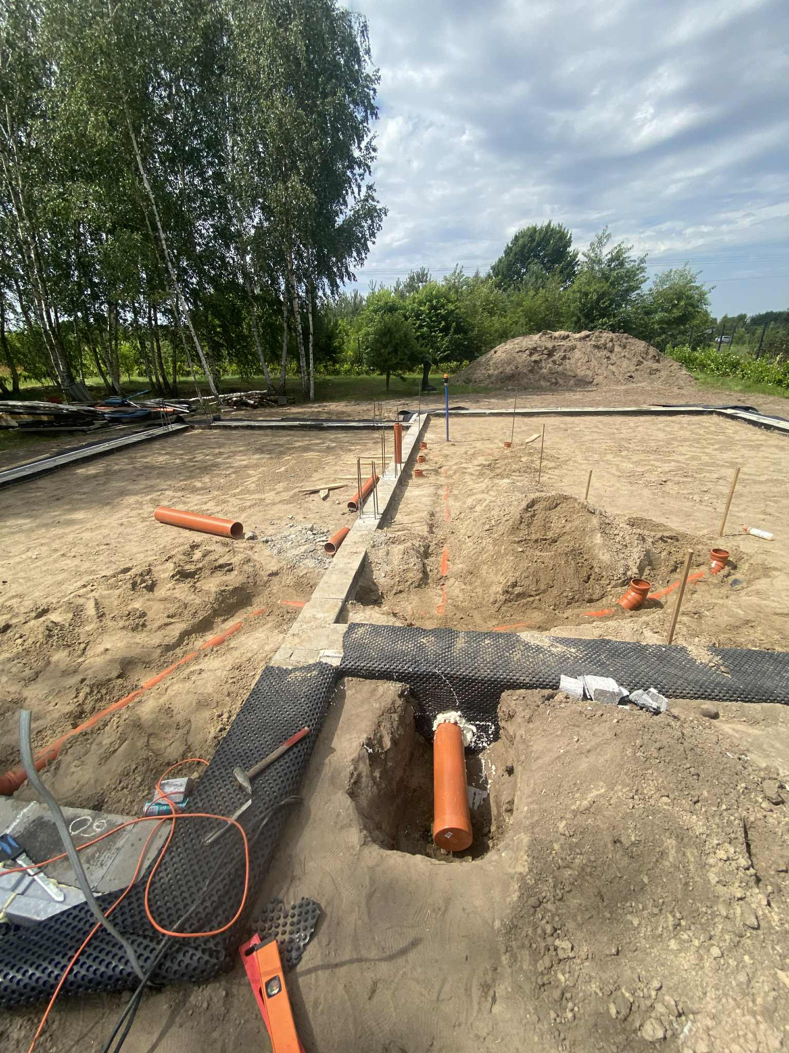 Fundament w trakcie budowy z widocznymi rurami kanalizacyjnymi, poziomica i narzędzia. W tle drzewa i usypana ziemia.