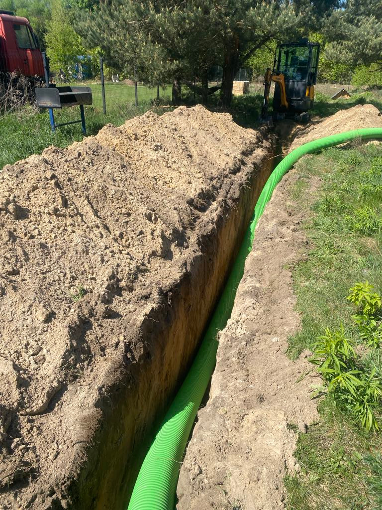 Wykopany rów z zieloną rurą drenażową w ziemi, widoczny mały traktor i fragment czerwonego samochodu. Słoneczny dzień, zielona trawa.