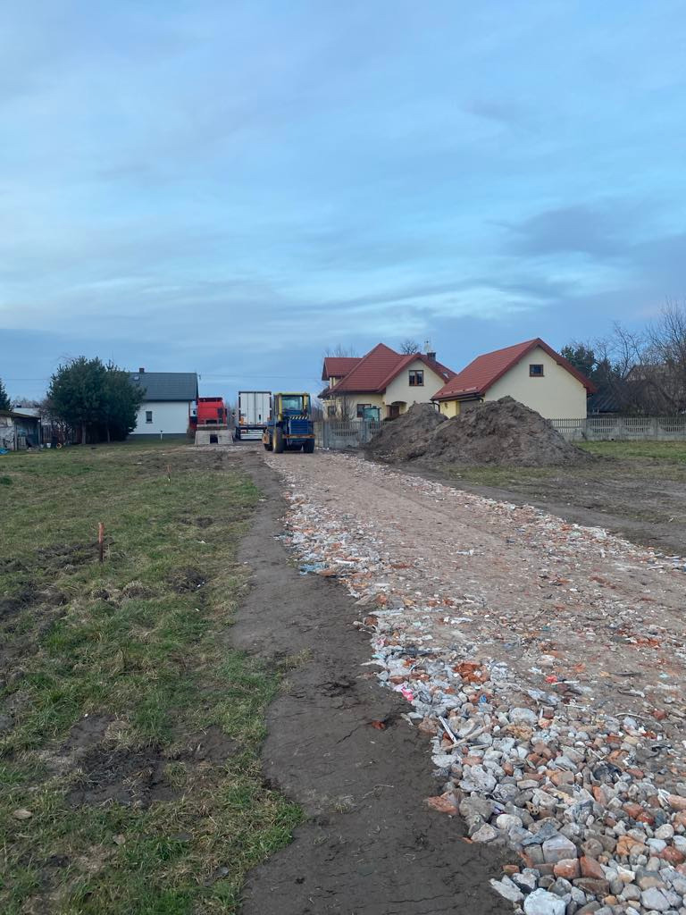 Wyrównywanie terenu gruzem i ziemią pod budowę w Słubicy A, z widocznym traktorem i stertą ziemi w tle domów. Prace ziemne przygotowawcze.