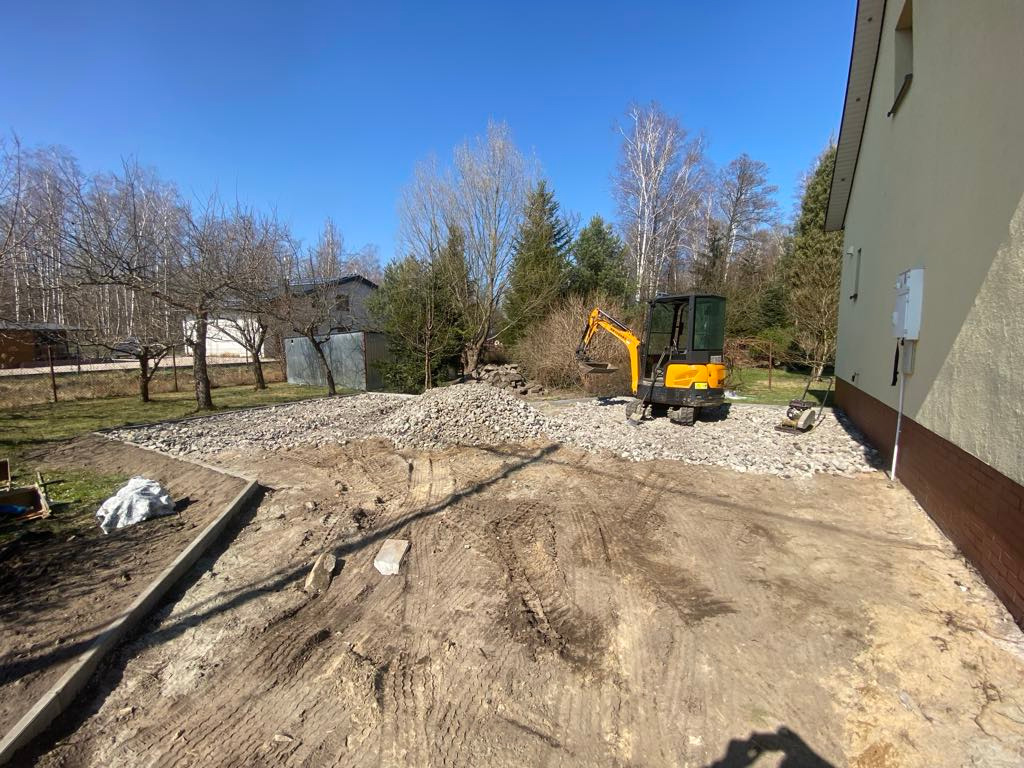 Żółta mini koparka na placu budowy, przygotowanie terenu pod kostkę brukową. Widoczne ślady opon i kamienie. W tle budynek i drzewa pod błękitnym niebem.