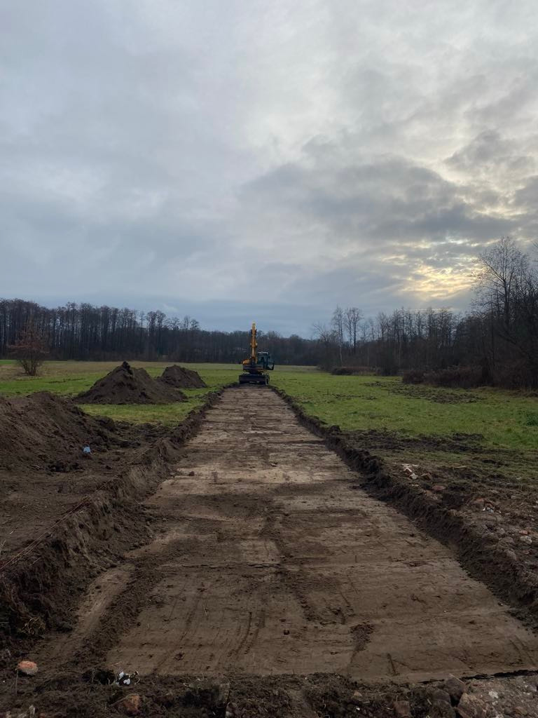Wykopany pas ziemi na polu, z koparką w oddali i stertami ziemi po bokach. Widok na horyzont pod pochmurnym niebem, sugerujący przygotowanie terenu pod budowę.