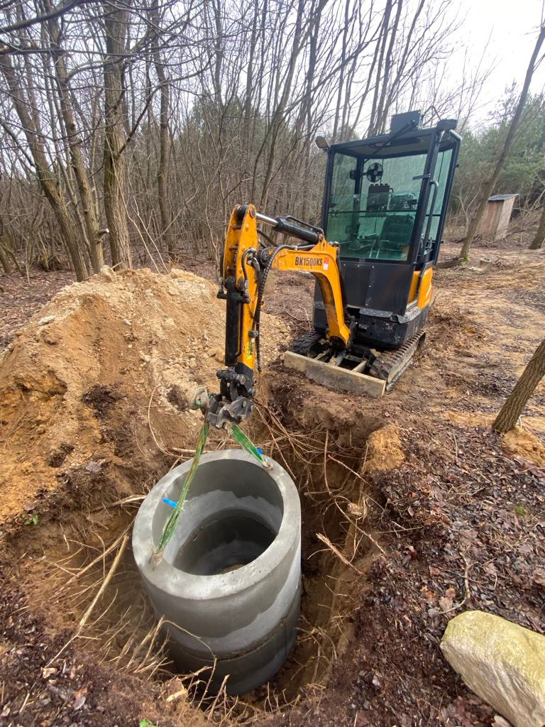 Żółta mini koparka BK1500KS opuszcza betonowy krąg do wykopu w ziemi, w tle las i mała drewniana budka. Widoczne korzenie i fragment kamienia.