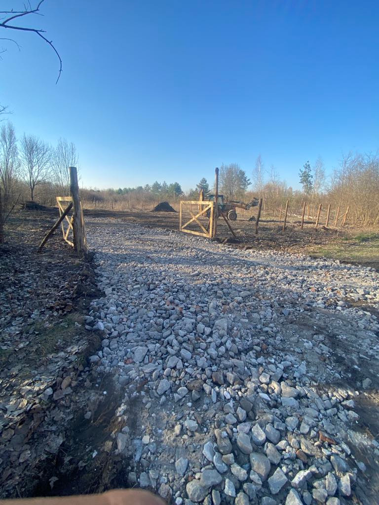 Ułożona droga z kamieni prowadząca przez drewnianą bramę na tle pola i lasu w słoneczny dzień. Widoczna maszyna budowlana w tle.