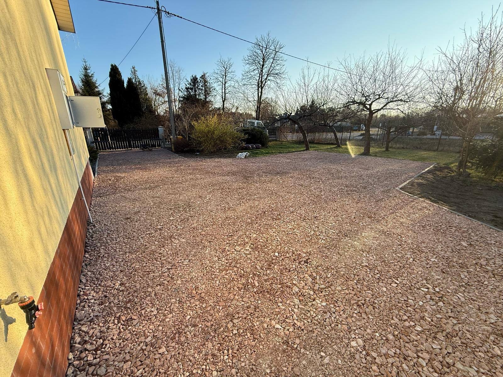 Utwardzony żwirem podjazd przy domu w słoneczny dzień. Widoczny kran na ścianie i granica trawnika. Żwir w odcieniach różu.