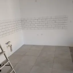 Świeżo wykafelkowana kuchnia z białymi płytkami typu metro na ścianie i szarymi płytkami podłogowymi, widoczne instalacje wodne i drabina.