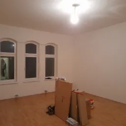 Pusty pokój przed malowaniem: widoczne trzy okna z zaokrąglonymi górnymi krawędziami, drewniana podłoga, białe ściany, kartony z narzędziami i farbami, oraz grzejnik.