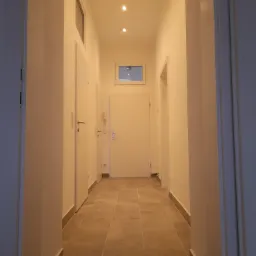 Długi, jasny korytarz z beżowymi płytkami podłogowymi, białymi ścianami i drzwiami. Widoczne listwy przypodłogowe w kolorze podłogi. Sufit z wpuszczonymi punktowymi lampami. Małe okno w górnej...