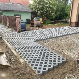 Ułożona ażurowa kostka betonowa na podjeździe w Niedzicy, widoczny fragment koparki, ogrodzenie z ceglanymi słupkami i budynek gospodarczy w tle.