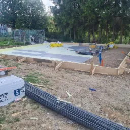 Przygotowanie podłoża pod fundament: drewniany szalunek, izolacja termiczna XPS, wiązka prętów zbrojeniowych na placu budowy w otoczeniu zieleni.