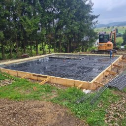 Heavy Solutions - Przygotowanie fundamentów pod budynek: deskowanie z drewnianych belek, siatka zbrojeniowa, w tle koparka na nierównym terenie z drzewami.