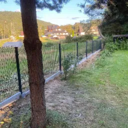 Nowo postawione, czarne, metalowe ogrodzenie panelowe z betonową podmurówką, widziane zza pnia drzewa na tle zabudowań wiejskich i zalesionych wzgórz.