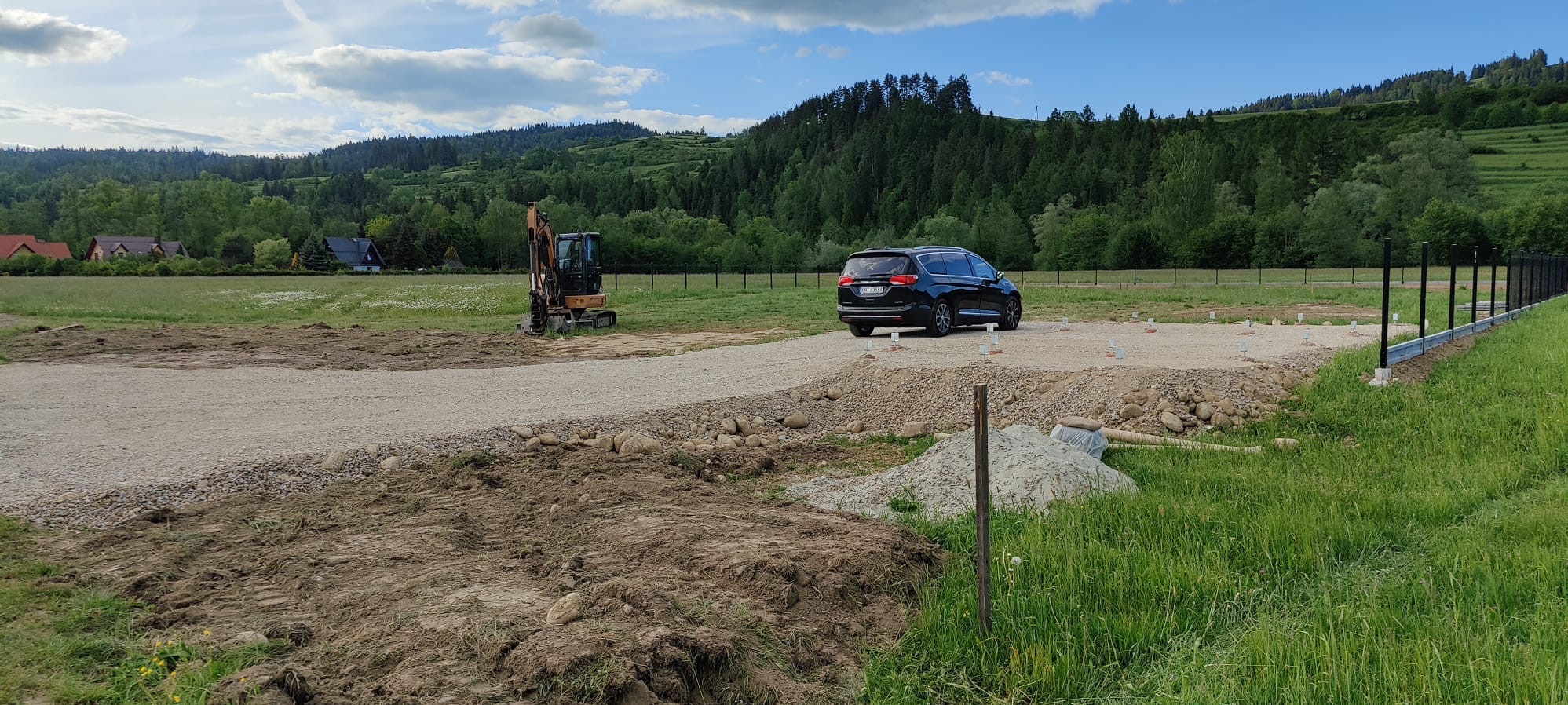 Przygotowany teren pod budowę z wytyczonymi punktami, częściowo utwardzony żwirem, z koparką w tle i zaparkowanym ciemnym vanem, ogrodzenie wzdłuż prawej granicy działki.