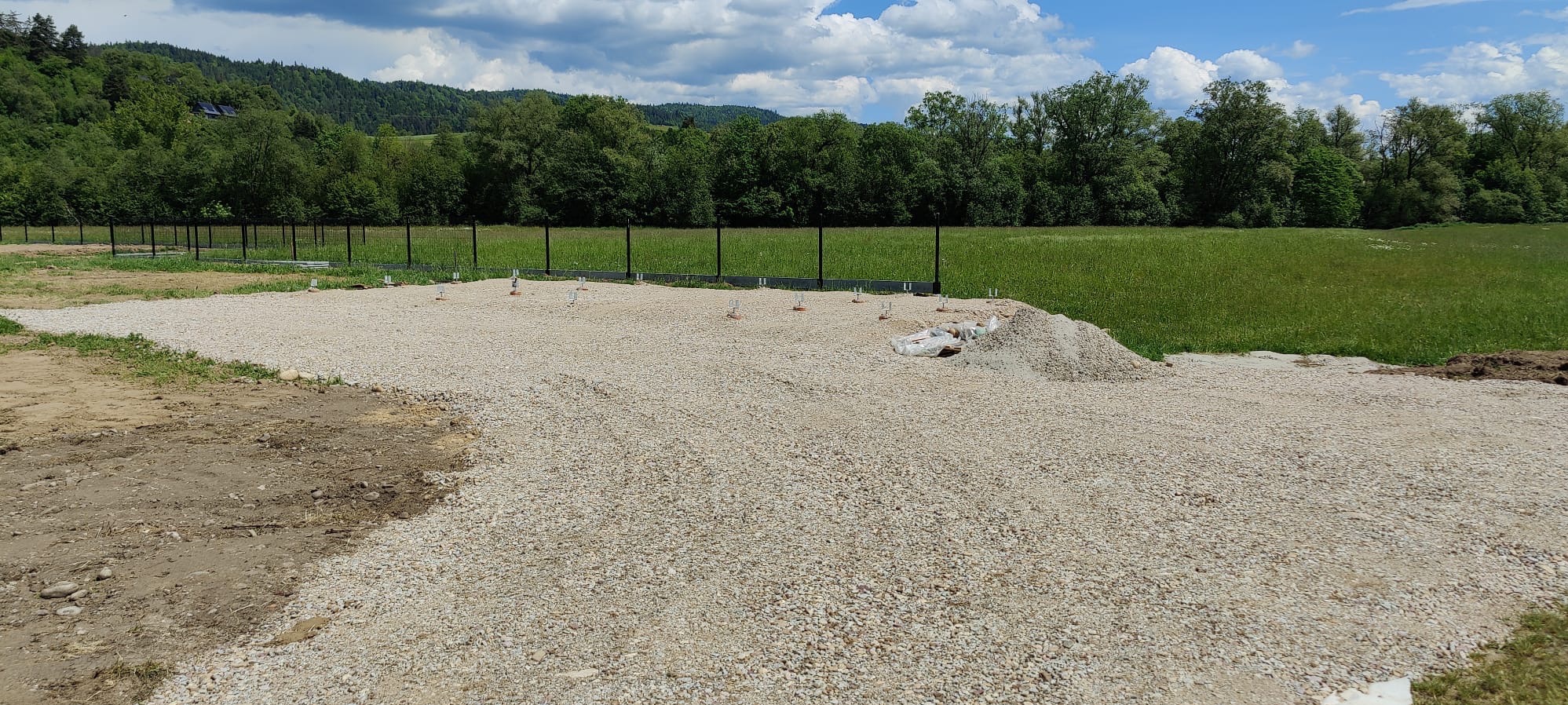 Przygotowany teren pod budowę z wysypanym żwirem, widoczne słupki ogrodzeniowe i fragmenty ogrodzenia w tle, teren otoczony zielenią.