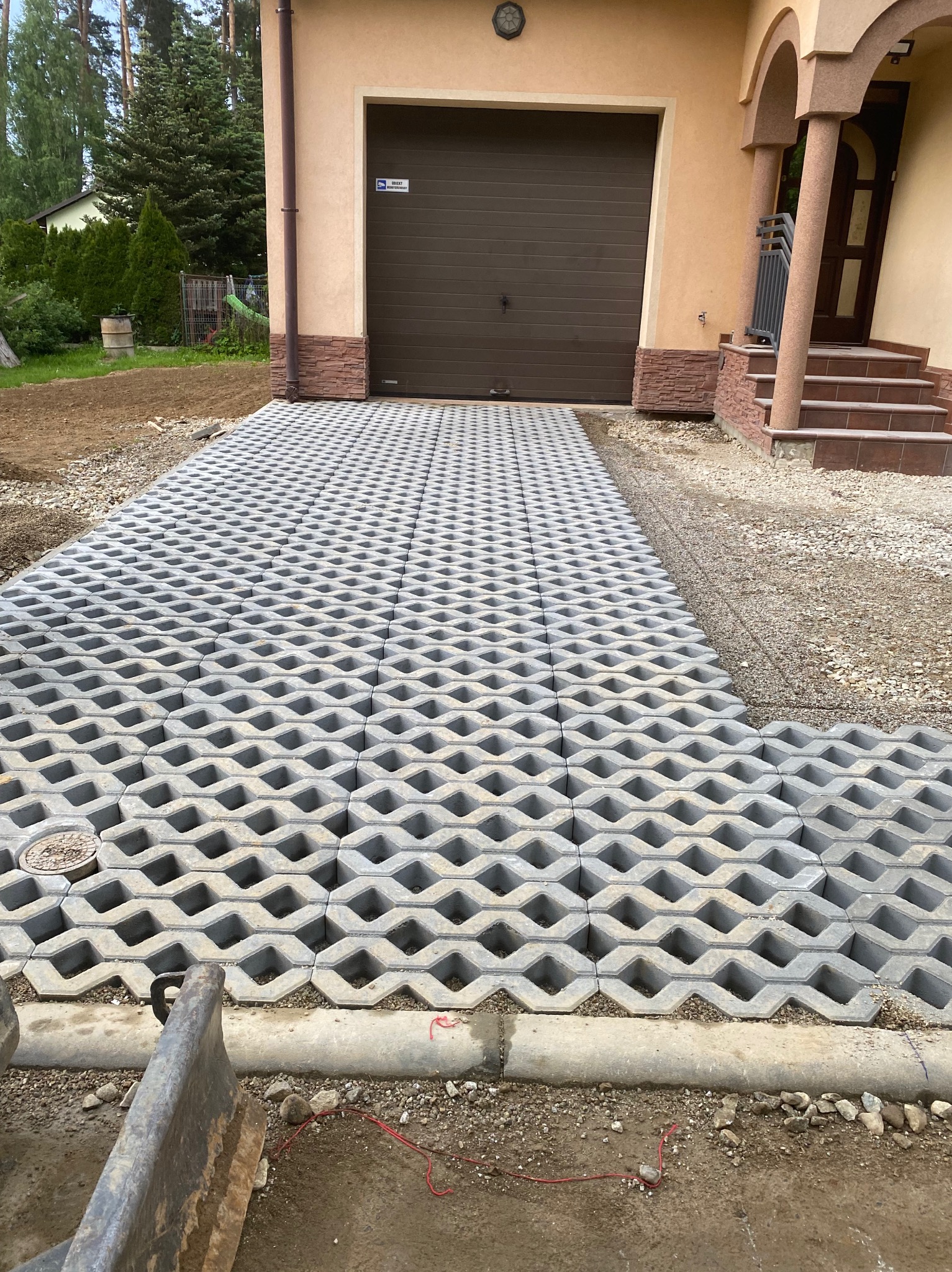 Niedokończony podjazd z ażurowych betonowych płyt w kształcie fal, z widocznymi śladami piasku i gruzu, prowadzący do garażu z brązową bramą segmentową i schodami wejściowymi do domu.